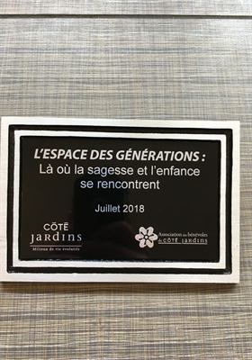Plaque noire encadrée de bois blanc, gravée du nom et de la date d’inauguration d’un espace intergénérationnel